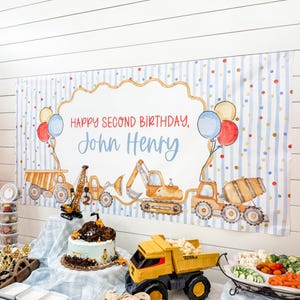 May include: A rectangular party banner featuring a light blue striped background with colorful dots. The center displays the text "HAPPY SECOND BIRTHDAY, John Henry" in red and blue, surrounded by watercolor illustrations of construction vehicles.