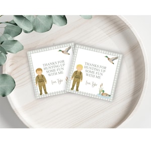 May include: Two white favor tags with a checkered border. Each tag features a cartoon illustration of a boy wearing camouflage clothing and a duck. The text on the tags reads "Thanks for hunting up some fun with me. Love. Tyler."