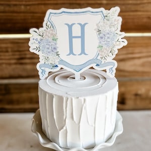 May include: A white cake with a blue and white floral cake topper with the letter H in the center. The cake is on a white cake stand.