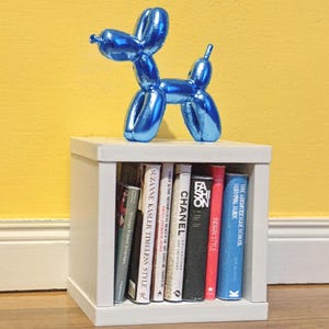May include: A shiny blue balloon dog sculpture sits atop a white cube shelf filled with books. The books have various colored spines and titles, including "CHANEL" and "INDIAN STYLE". The background is a solid yellow.