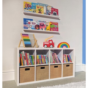 May include: A white bookshelf unit with books, baskets, and toys. The unit has cubby holes with books and woven baskets. Above are two white shelves with children's books. A red fire truck toy and a rainbow are on top.