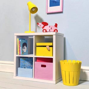 May include: A white cube shelf unit with four compartments, holding books, boxes, and a yellow lamp. A pink dog toy and a yellow wastebasket are on the shelf. The boxes are blue, pink, and yellow.