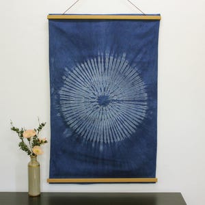 May include: A rectangular indigo blue fabric wall hanging featuring a central white sunburst tie-dye pattern. The textile is secured between two horizontal light-colored wooden slats at the top and bottom, suspended by a thin dark string.