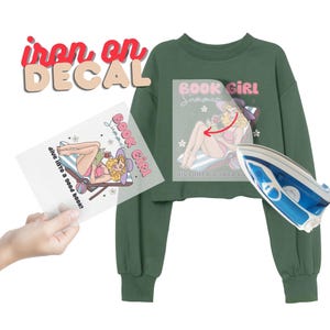 May include: An iron-on decal with the words "iron on DECAL" in red and pink. The decal features a cartoon illustration of a woman in a swimsuit reading a book, with the words "BOOK GIRL Summer" and "dive into a good book!" printed on it. A green sweatshirt and iron are also in the image.