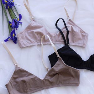 May include: Three triangle bras in neutral colors: light pink, black, and taupe. Each bra has adjustable straps and a simple design. The bras are arranged on a white, textured surface, with a sprig of purple irises in the upper left corner.