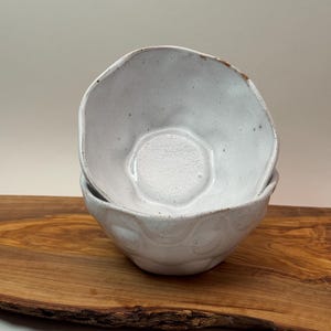 May include: Two white ceramic bowls with irregular edges are stacked on a wooden board. The bowls have a speckled texture and a slightly uneven surface. The bowls are a neutral colour and would be suitable for serving food.