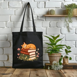 Puede incluir: Bolso tote negro con un diseño de un gato atigrado naranja durmiendo sobre una pila de libros. Los libros son rojos, verdes y marrones, rodeados de flores y vegetación. El bolso tiene un asa larga.
