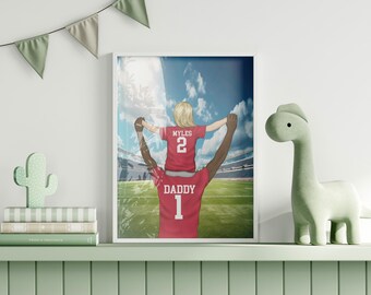 Op de afbeelding: Een ingelijste kunstdruk met een persoon die een kind op de schouders draagt, beiden in rode shirts met witte letters. Het shirt van het kind toont MYLES 2 en dat van de volwassene DADDY 1, tegen de achtergrond van een voetbalstadion.