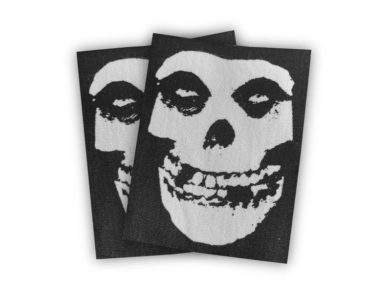 May include: Two black fabric patches with a white smiling skull design. The skull has two eye sockets, a nose, and a wide, toothy grin.
