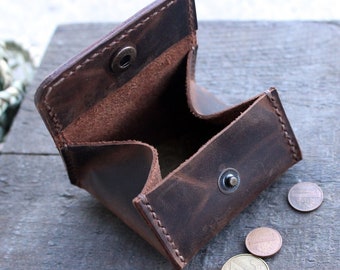 May include: Open, brown leather coin purse with a snap closure. The purse is empty, with a few coins scattered nearby. The leather has a distressed look, and the stitching is visible. The purse is on a wooden surface.