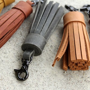 May include: Close-up of several leather tassels in various colors, including tan, brown, and gray. Each tassel is attached to a metal clasp. The tassels are made of strips of leather and are likely used as keychains or decorative accessories.