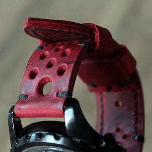 以下が含まれることがあります： 黒い文字盤が付いた赤い革の時計ストラップのクローズアップ。ストラップには複数の円形の穴と黒いステッチがあります。文字盤は黒で、白い数字が付いています。