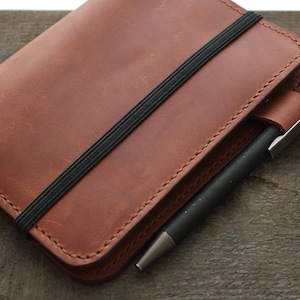 May include: A brown leather notebook cover with a black elastic band closure. A black pen is inserted into a leather pen holder on the side. The notebook cover is made of a smooth, textured leather.