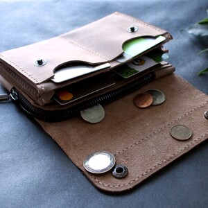 May include: Open, light brown leather wallet with a zipper closure and snap button details. The wallet is partially open, revealing credit cards and coins. The interior is a lighter shade of brown.