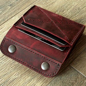Può includere: Un portafoglio in pelle bordeaux con chiusura a scatto e cuciture a vista. Il portafoglio è aperto, rivelando diverse carte di credito. La pelle ha un aspetto invecchiato e il portafoglio è su una superficie di legno.