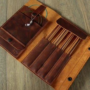 May include: Open leather knitting needle case in shades of brown. The case is open, revealing pockets for needles and accessories. The case is made of distressed leather and has a button closure. The case contains knitting needles and other tools.