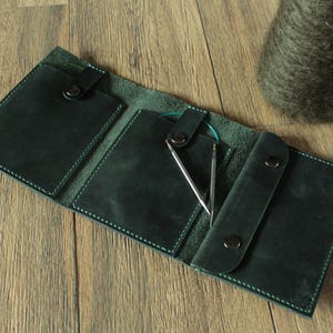 May include: Dark green leather knitting needle case, open to display needles and a circular needle. The case has multiple pockets and a snap closure. A ball of yarn is in the background. The case is on a wooden surface.