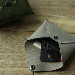 May include: A gray leather card holder, open to reveal cards and cash. The triangular wallet is secured with a snap closure. A second, olive green wallet is visible in the background. The wallets are on a wooden surface.