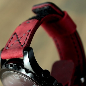May include: Close-up of a black wristwatch with a red leather strap. The strap is stitched with black thread and has a textured appearance. The watch face is visible, with white numerals and hands.
