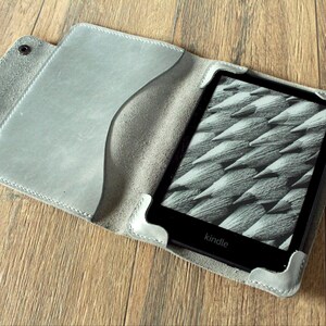 Könnte beinhalten: Eine graue Kindle-Hülle aus Leder, geöffnet, um den E-Reader anzuzeigen. Die Hülle hat einen Druckknopfverschluss und eine Tasche. Der Kindle-Bildschirm zeigt ein Schwarz-Weiß-Bild. Die Hülle liegt auf einer Holzoberfläche.