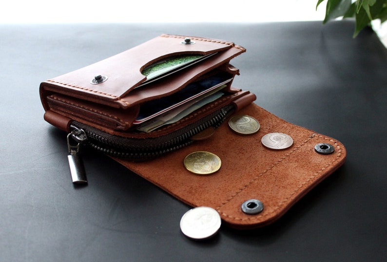 May include: A brown leather wallet with a zipper closure and snap button. The wallet is open, revealing cards and coins. The wallet is made of brown leather and has a silver zipper.