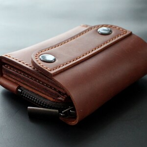 May include: A brown leather wallet with a zipper closure and snap closure. The wallet is made of a rich, textured leather and features detailed stitching. The wallet is designed to hold cards and cash.
