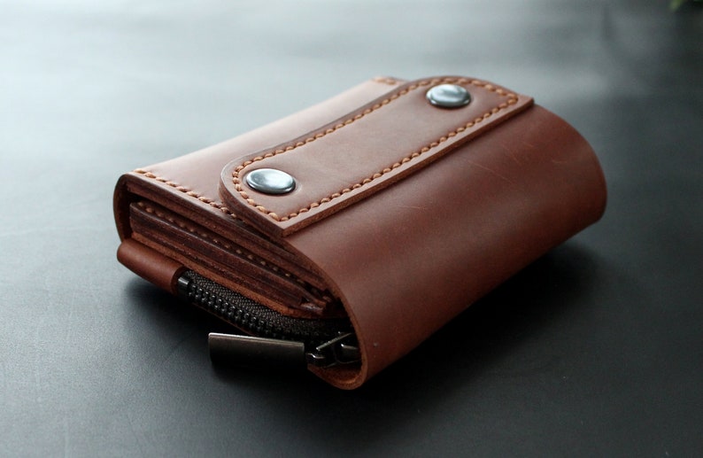 May include: A brown leather wallet with a zipper closure and snap closure. The wallet is made of a rich, textured leather and features detailed stitching. The wallet is designed to hold cards and cash.