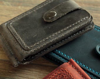 May include: Close-up of a gray leather money clip with a snap closure. The clip is stitched with a contrasting thread and has a worn, textured appearance. Other leather wallets in teal and red are visible.