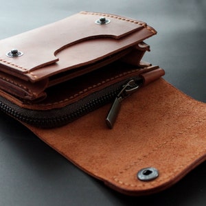 May include: Open brown leather wallet with a zipper closure and snap button details. The wallet is partially open, revealing interior compartments. The leather has a natural grain and texture, with visible stitching.