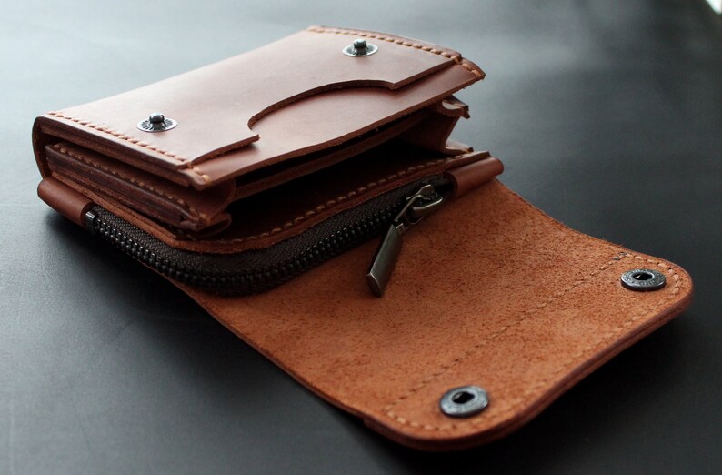 May include: Open brown leather wallet with a zipper closure and snap button details. The wallet is partially open, revealing interior compartments. The leather has a natural grain and texture, with visible stitching.