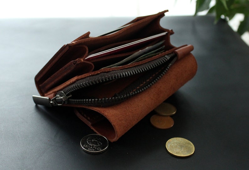 May include: A brown leather wallet with a zipper closure, open to reveal cards and cash. Several coins are scattered near the wallet. The wallet is made of a textured, suede-like material.