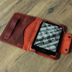 May include: Open leather Kindle case in a reddish-brown hue. The case features a handle, a snap closure, and a built-in stand. The Kindle device is visible inside the case, displaying a newspaper-like image on its screen.