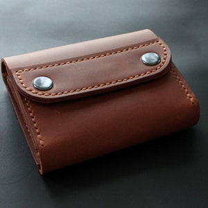 May include: A brown leather wallet with a flap closure secured by two silver-colored snap buttons. The wallet features visible stitching in a lighter brown thread. The leather has a smooth texture.