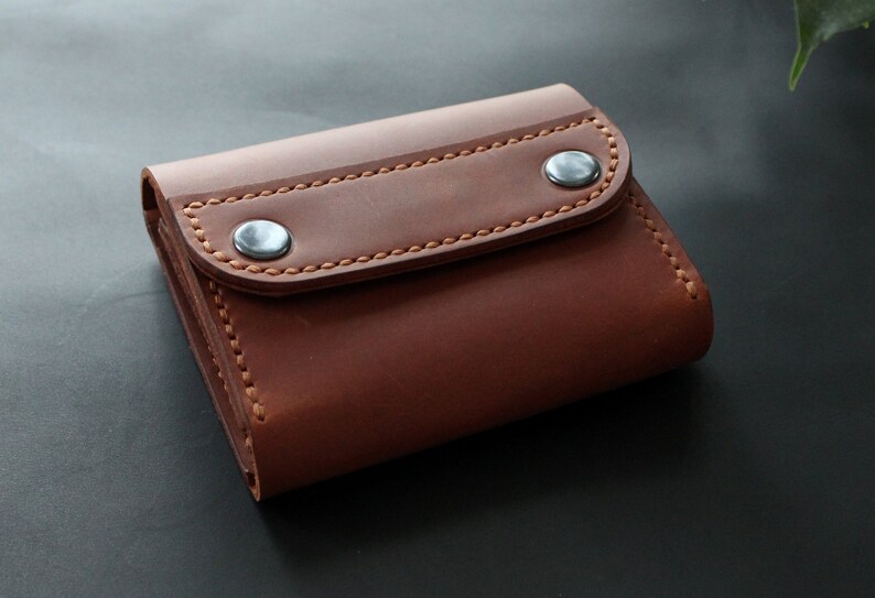 May include: A brown leather wallet with a flap closure secured by two silver-colored snap buttons. The wallet features visible stitching in a lighter brown thread. The leather has a smooth texture.