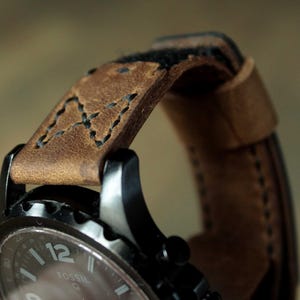May include: Close-up of a Fossil watch with a brown leather strap. The watch has a black face with white numerals and hands. The leather strap is stitched with black thread and shows wear.