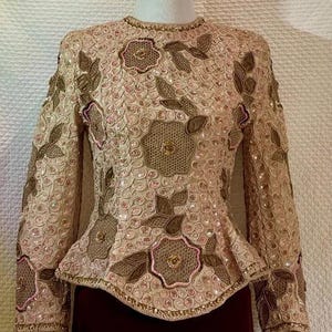 May include: A vintage, long-sleeved top in a beige hue, adorned with intricate floral embroidery and sequin embellishments. The design features brown and pink floral patterns, a beaded neckline, and a peplum waist. The top is displayed on a mannequin.