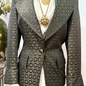 May include: A dark gray blazer featuring a repeating geometric pattern in a metallic gold tone. The blazer has a single front button, notched lapels, and cuffed sleeves. It is worn over a white collared shirt with a layered gold chain necklace.