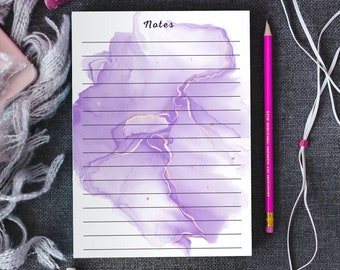 Puede incluir: Un bloc de notas con líneas y un diseño de acuarela púrpura con la palabra "Notes" en la parte superior. El bloc de notas está sobre una superficie gris con un teléfono rosa, un lápiz rosa y un par de auriculares rosas.
