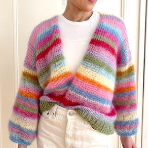 Könnte beinhalten: Ein farbenfroher gestreifter Cardigan mit V-Ausschnitt und langen Ärmeln. Der Cardigan ist aus weichem, flauschigem Garn gefertigt und in verschiedenen Farben gehalten, darunter Rosa, Blau, Grün, Gelb und Orange.