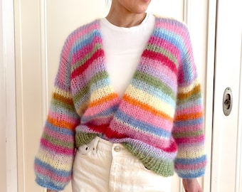 May include: A colorful, open-front cardigan with horizontal stripes in shades of pink, blue, yellow, orange, and green. The sleeves are long and the knitwear has a fluffy texture. The cardigan is worn over a white top.