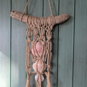 Può includere: Un arazzo da parete in macramè con un ramo di legno, corda naturale e conchiglie. La corda beige è annodata per creare un motivo decorativo, con tre conchiglie rosa e bianche integrate nel design. L'arazzo è sospeso da una corda.