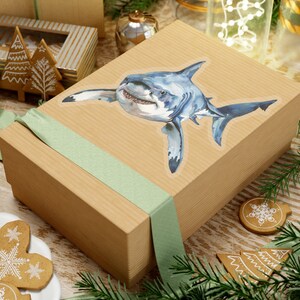 May include: A brown gift box with a green ribbon and a white shark sticker. The shark is facing the viewer with its mouth open.