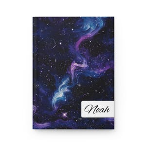 Op de afbeelding: Een donkerblauw notitieboek met een galaxy-print met draaiende paarse en blauwe nevels en een strooiing van witte sterren. Het notitieboek heeft een wit label met de naam "Noah" in zwart gedrukt.