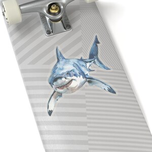 May include: A white skateboard deck with a gray and white striped pattern. A watercolor illustration of a great white shark is on the deck. The shark is swimming towards the right side of the image.
