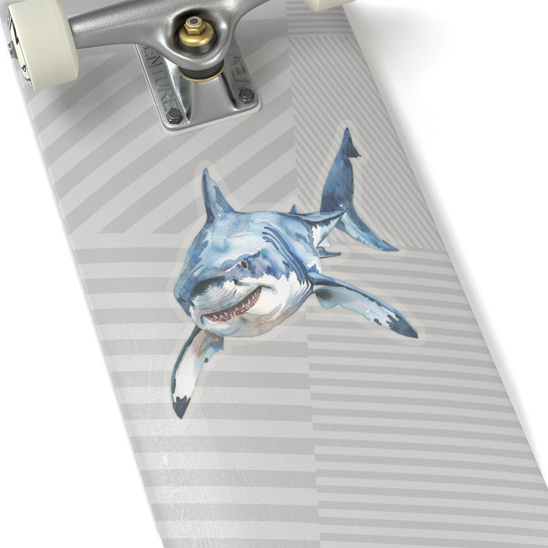 May include: A white skateboard deck with a gray and white striped pattern. A watercolor illustration of a great white shark is on the deck. The shark is swimming towards the right side of the image.
