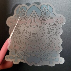 May include: A holographic sticker with a detailed, symmetrical design. The sticker features a complex pattern of lines and shapes, with a color gradient of blue and pink. The sticker is held by a pink object.