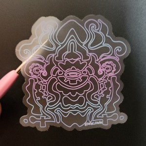May include: A clear sticker with a purple and blue outline of a complex geometric design. The design is reminiscent of a face with a large eye and a mouth. The sticker has the text "@Parker.Crafts" in the bottom right corner.