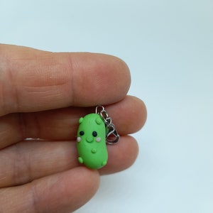 Op de afbeelding: Een groene augurkenhanger met een glimlachend gezicht, witte ogen en zwarte pupillen. De hanger is bevestigd aan een zilveren metalen ketting.
