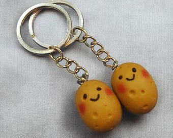 Puede incluir: Dos llaveros con forma de patata con caras sonrientes y mejillas rosadas. Los llaveros están conectados por una cadena dorada y sujetos a un llavero plateado. Las patatas son de color marrón claro.