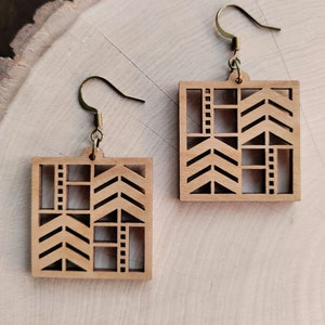 Puede incluir: Un par de pendientes cuadrados de madera con un diseño geométrico. Los pendientes son de color marrón claro y cuelgan de ganchos dorados. El diseño incluye flechas y cortes rectangulares.